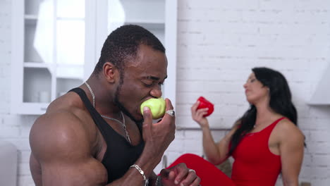 african man and caucasian woman eating fresh fruit. black skinned sportive guy eating apple next to the beautiful woman in red dress in the kitchen.