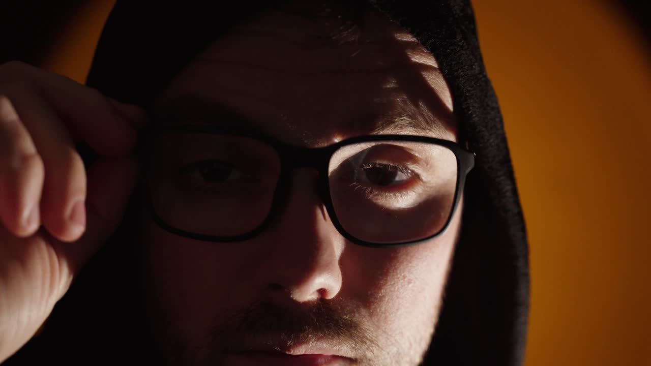 Primer plano cinematográfico de un hombre serio con sudadera con capucha y gafas bajo una luz baja dramática, creando una atmósfera misteriosa e intensa a cámara lenta.