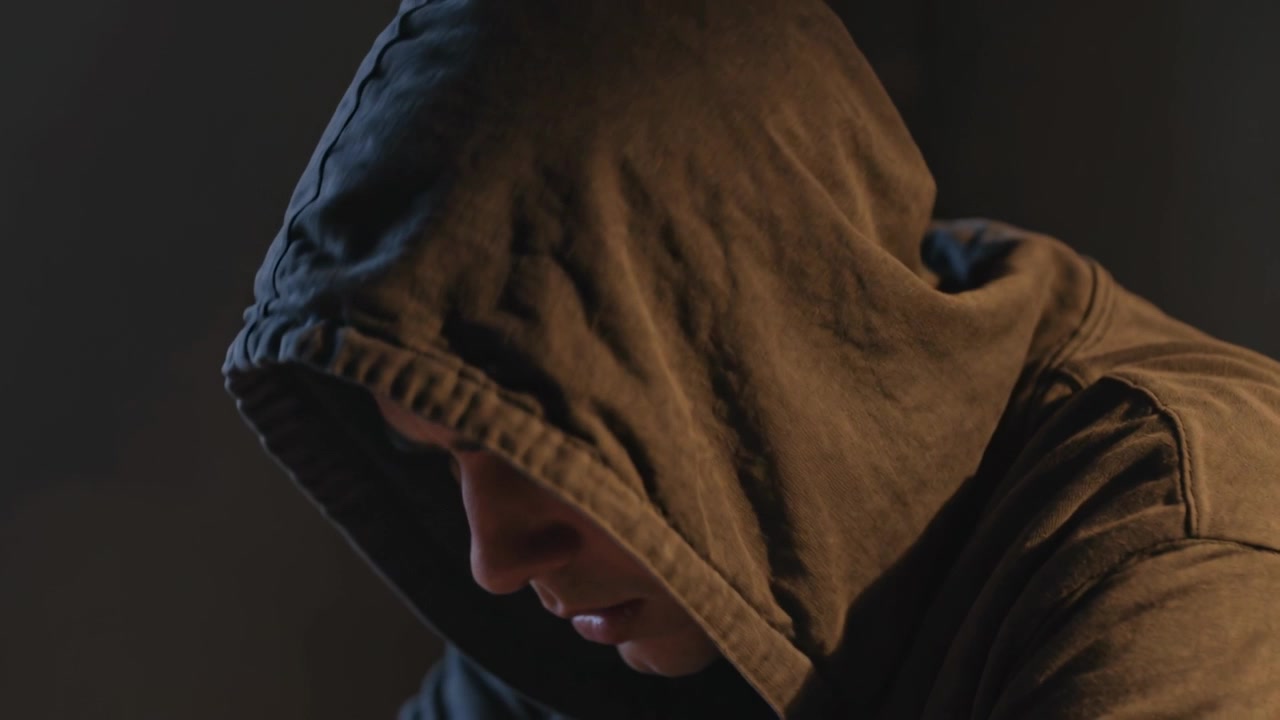 Sekansın başlangıcına tepki veren, loş bir odada oturan adam çenesini indirip konuşuyor, üzerinde kapüşonlu bir sweatshirt var.