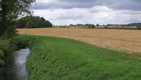 Tractor-in-the-distance-working-the-land-in-a-beautiful-field,-with-a-running-stream-in-the-foreground