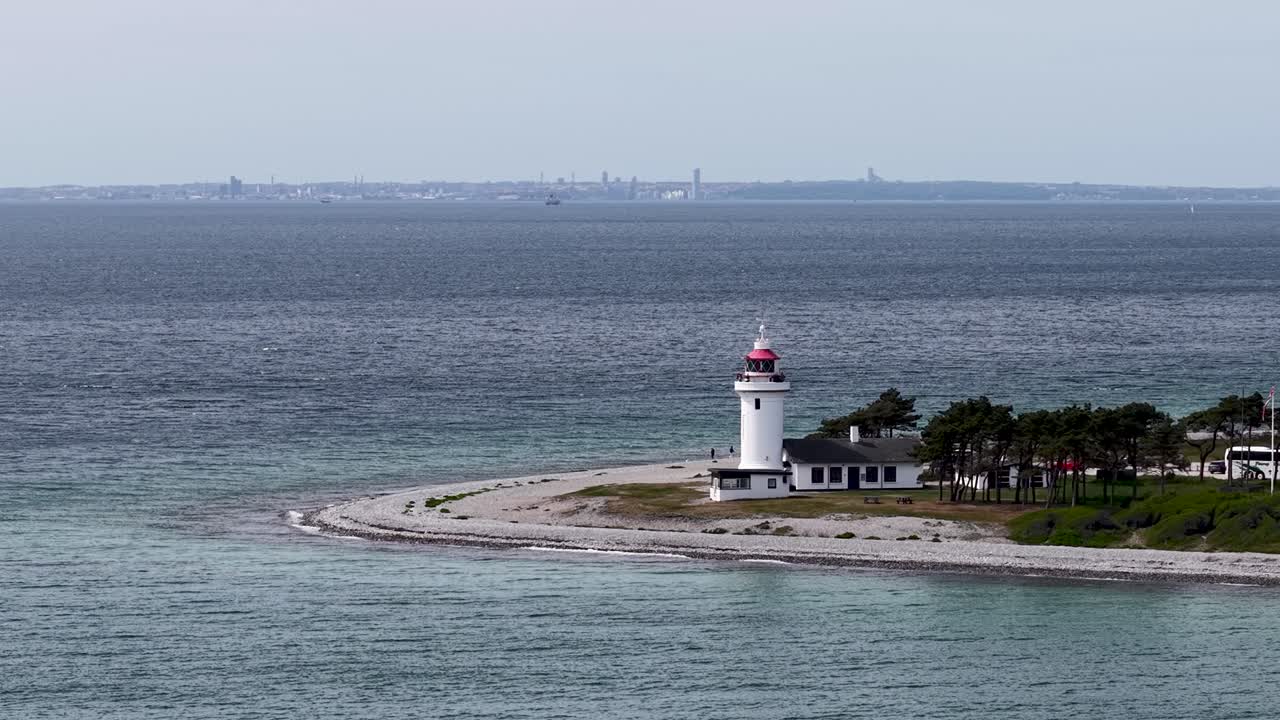 Aerial view of a small lighthouse perched on a narrow peninsula jutting into the sea, with gentle waves lapping the rocky shore and a distant city skyline across the bay