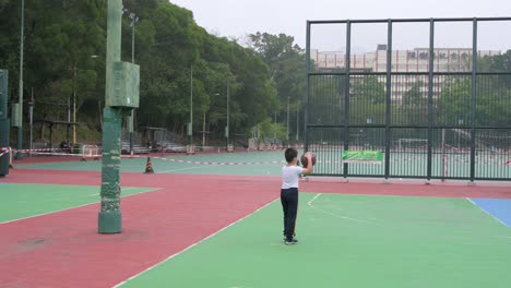 Ein-Kind,-Das-Eine-Gesichtsmaske-Trägt,-Während-Es-Einen-Basketballball-Hält,-Geht-Auf-Einem-Leeren,-Farbenfrohen-Basketballplatz-Spazieren,-Der-Wegen-Des-Ausbruchs-Und-Der-Beschränkungen-Des-Covid-19-Coronavirus-Geschlossen-Ist