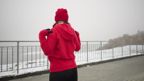 back view of coach in red hoodie outdoors performing neck stretches with hands on shoulders, turning head left and right in a serene winter setting with snow-covered ground and iron railing