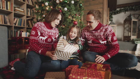 happy little girl unwrapping gift while sitting with her mom and dad in front of christmas tree at home 3