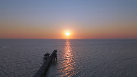 drone hovers still above the ocean, capturing majestic waves beneath a glowing sunset. the pier stretches into view while golden hues light the sky in a serene, powerful coastal moment.