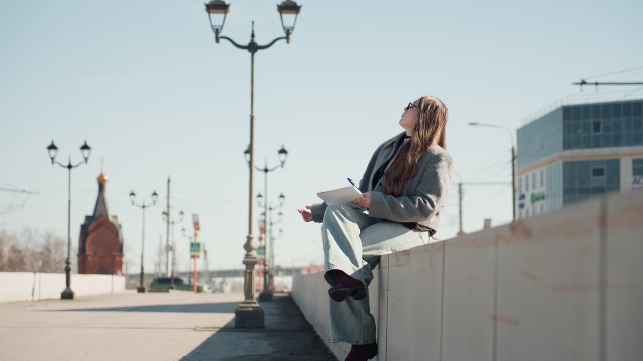 Ładna kaukaska projektantka przeglądająca notatki na zewnątrz, siedząca na betonowym gzymsie w pobliżu centrum handlowego, w zrelaksowanej pozie, z notatnikiem na kolanach, w miękkim świetle słonecznym i z panoramą miasta w tle, w optymistycznym, twórczym nastroju.
