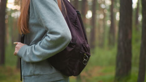 close-up of young woman hiking through forest wearing gray sweater and backpack, standing amidst lush greenery, focus on arm and hand grasping backpack strap