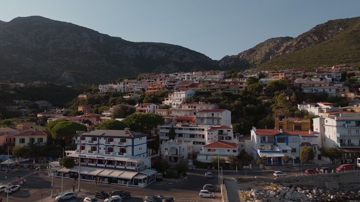 Perto do Porto de Cala Gonone, Sardenha, Itália, mostrando casas e edifícios à beira-mar sob colinas escarpadas sob a luz quente da tarde ao lado da costa do Mediterrâneo, vista aérea panorâmica
