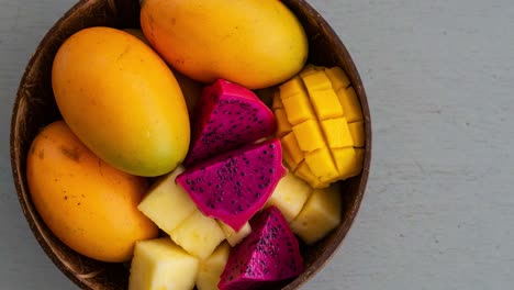 a vibrant display of tropical fruits: golden mangos, bright dragon fruit, and sweet pineapple arranged in a rustic bowl on a soft gray surface