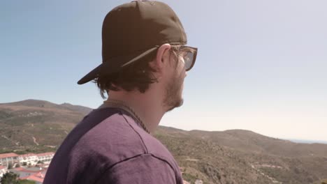 Man-with-baseball-hat-and-sunglasses-walking-around-hills-on-sunny-summer-day