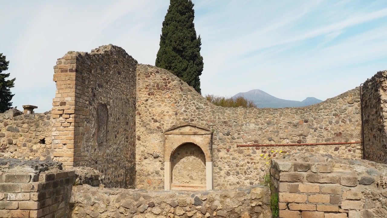 Antiche rovine di Pompei con muri in pietra consumati dal tempo, muratura in mattoni, piccola nicchia votiva, un cipresso e il Vesuvio sullo sfondo.