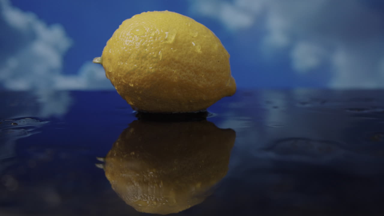Gocce di pioggia cadono su una superficie bagnata riflettente mentre un limone giallo rimane immobile e le nuvole si spostano in un cielo nuvoloso in time lapse