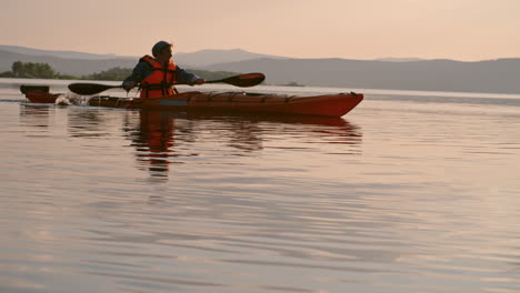 Seitenansicht-Eines-Mannes-Mit-Mütze-Und-Schwimmweste,-Der-Bei-Sonnenuntergang-Ein-Kanu-Auf-Dem-See-Paddelt