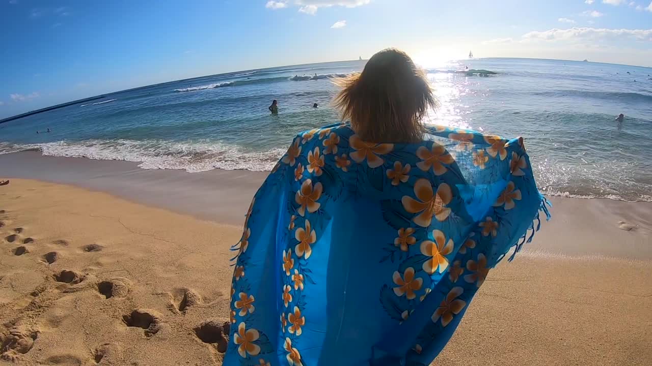 atractiva chica rubia caminando hacia el océano con sarong hawaiano sopla en el viento en cámara lenta