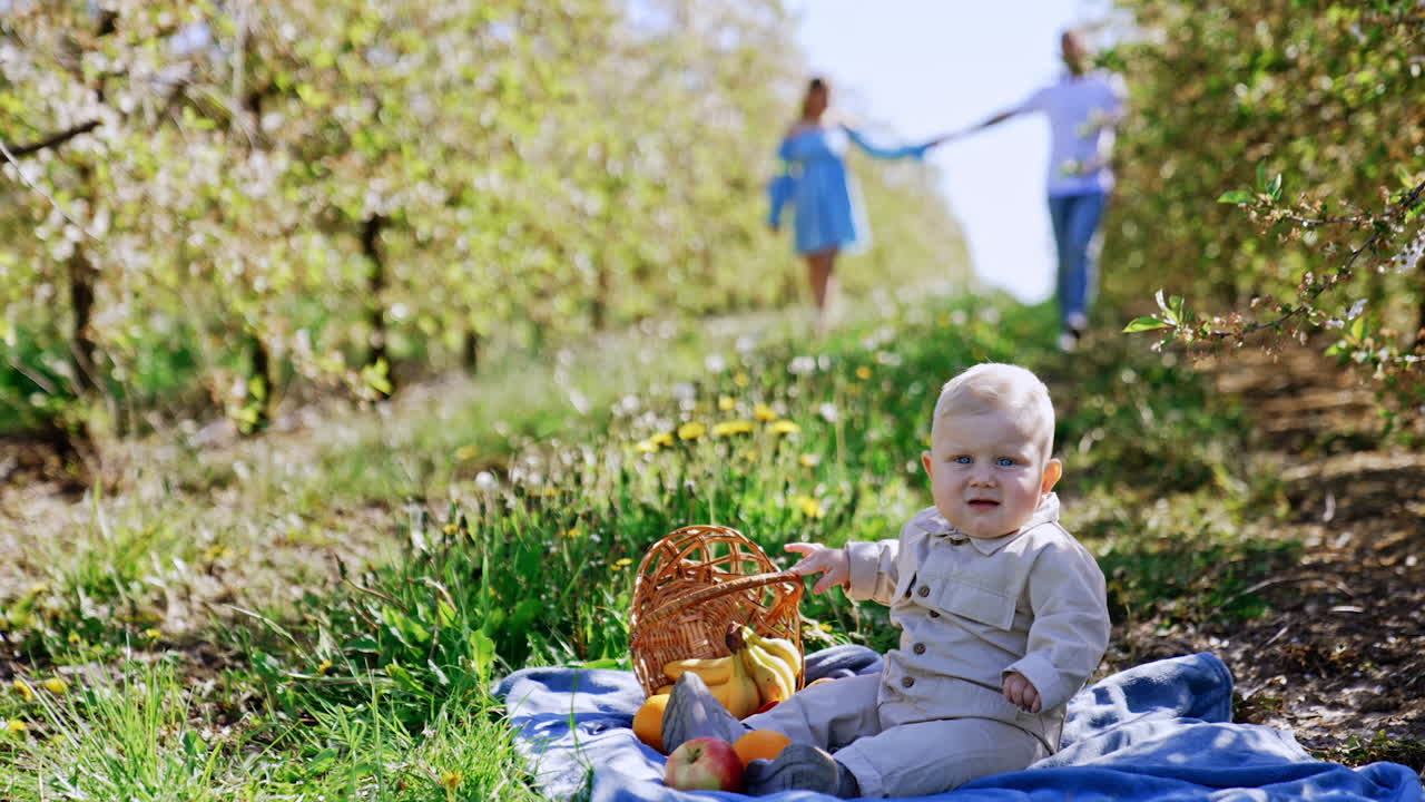 Adorable blond baby boy holding a fruit basket sits on the ground. Parents holding their hands walk by the garden at backdrop in blur.