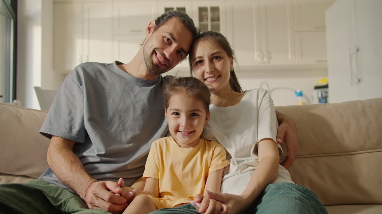 Portrait of a happy family, a brunette dad with stubble, his brunette girlfriend in a white T-shirt and their happy daughter in a yellow dress sitting on a light brown sofa in a modern studio apartment