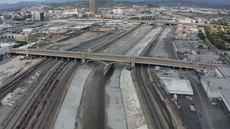 AERIAL:-Los-Angeles-River-Bridge-with-Tram-Train-Crossing-on-Cloudy-Overcast-Sky