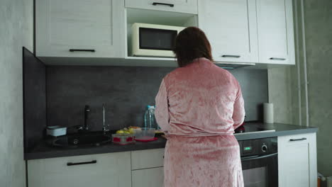 woman in pink velvet robe seen from behind standing at kitchen counter preparing food, surrounded by apples, bread, containers, and bottles, creating quiet morning domestic scene with calm natural light