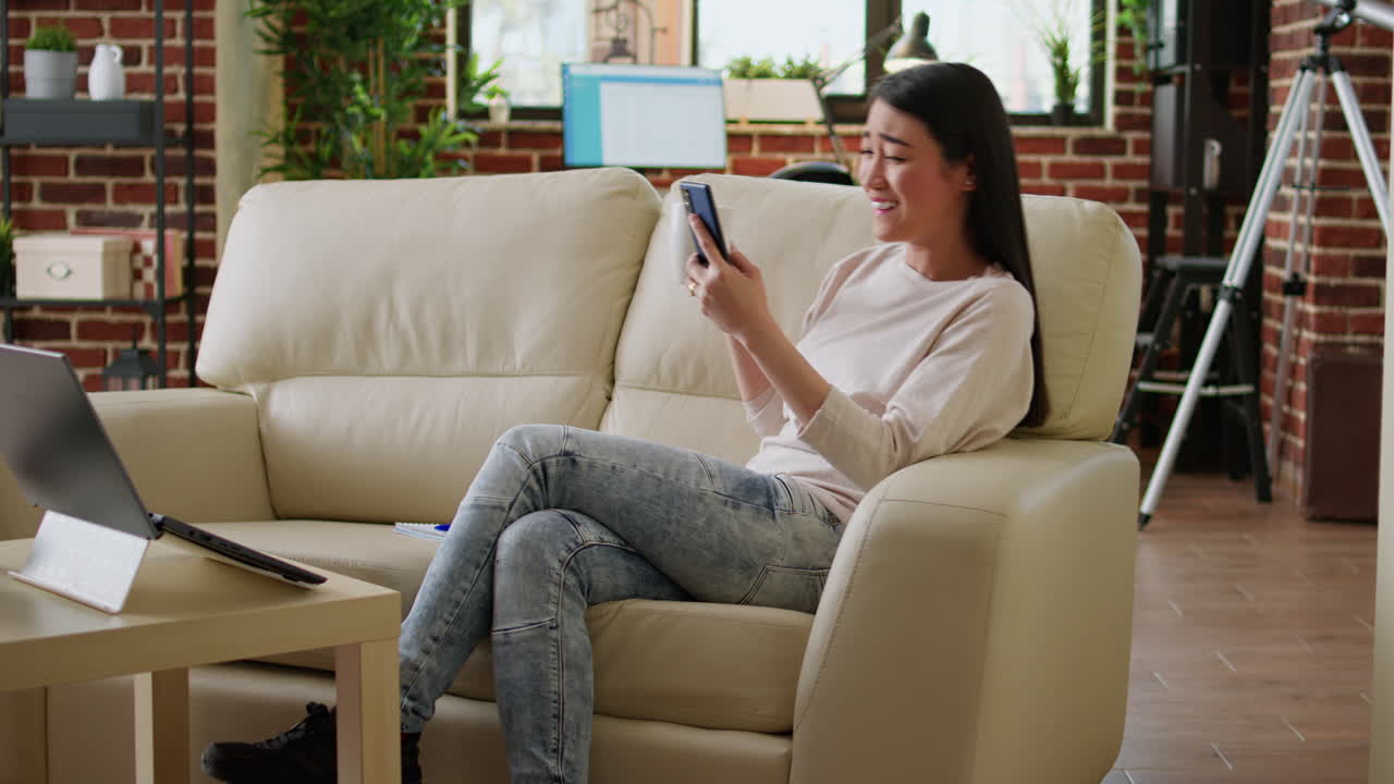 Woman using smartphone while relaxing on couch with laptop