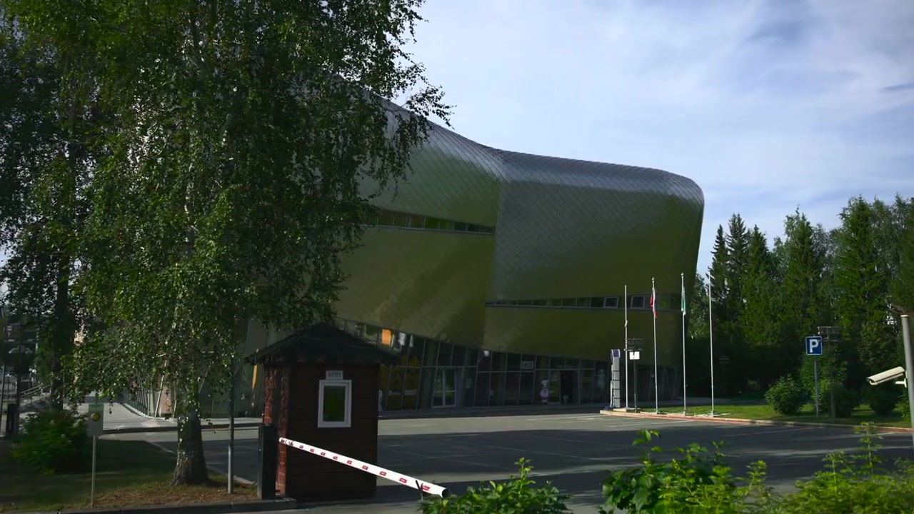 El brazo de la barrera se eleva desde la posición cerrada para abrir el acceso en una entrada cívica contemporánea junto a un edificio metálico curvado, con abedules, superficies de cristal, calzada, pavimento y una tranquila escena diurna