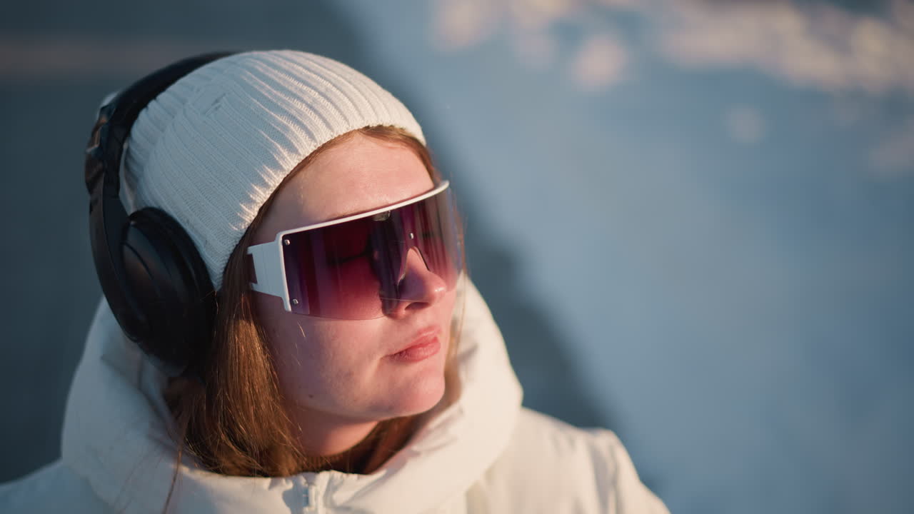 Close up shot of female artist wearing tinted goggles and white beanie smiling under sky with sun reflecting off visor as breeze tousles hair against snow backdrop evoking winter fashion mood