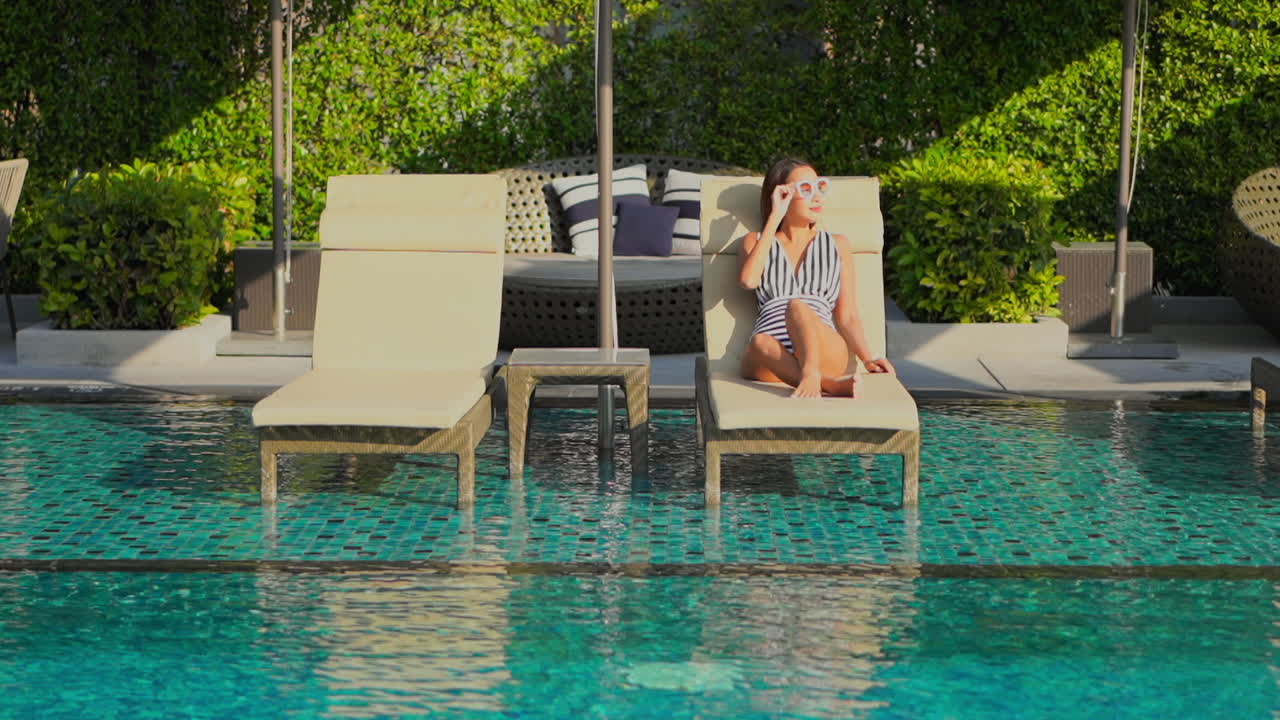 Sexy Asian woman wearing a striped swimming suit and sunglasses resting tanning on a poolside lounge deck chair at a luxury resort in Thailand