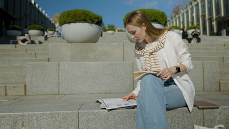 beautiful female college student sitting on stairs outdoor, writing notes on a notebook in university campus