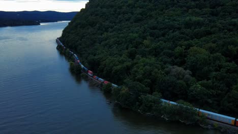 aerial drone footage of a long freight train going along the tracks at the base of a mountain next to a river in the appalachian mountains at sunset