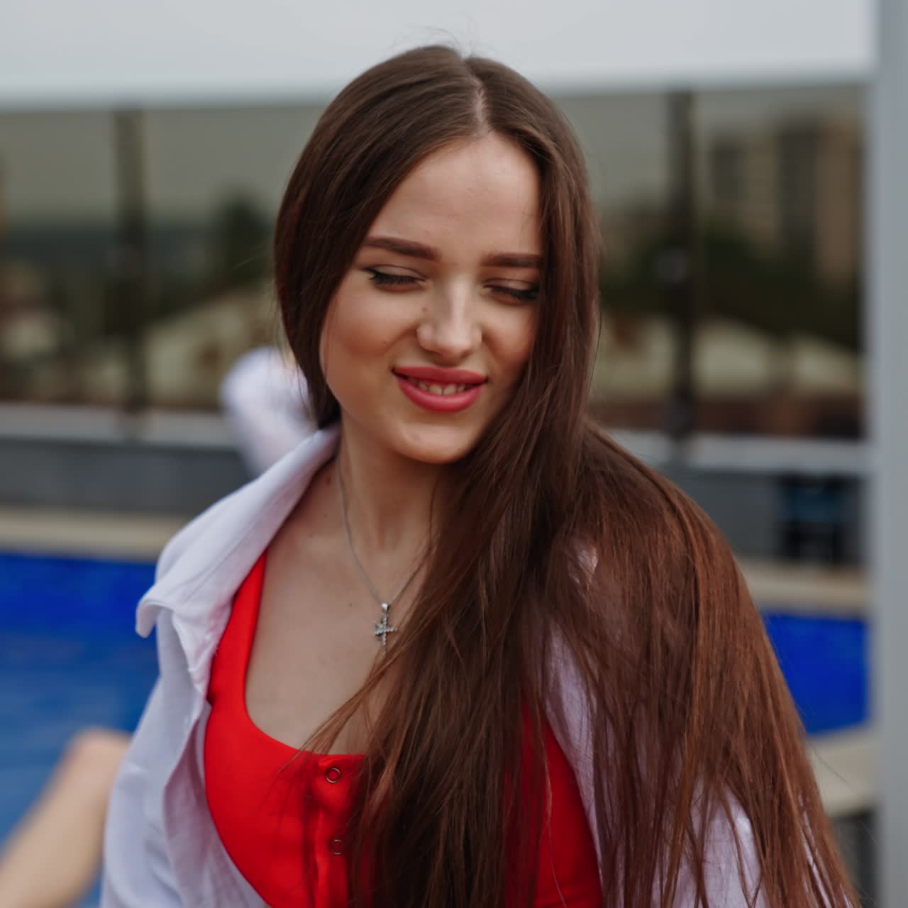 Beautiful brunette in red swimsuit and white shirt walks and turns in front of camera. Female model posing near the pool outdoors