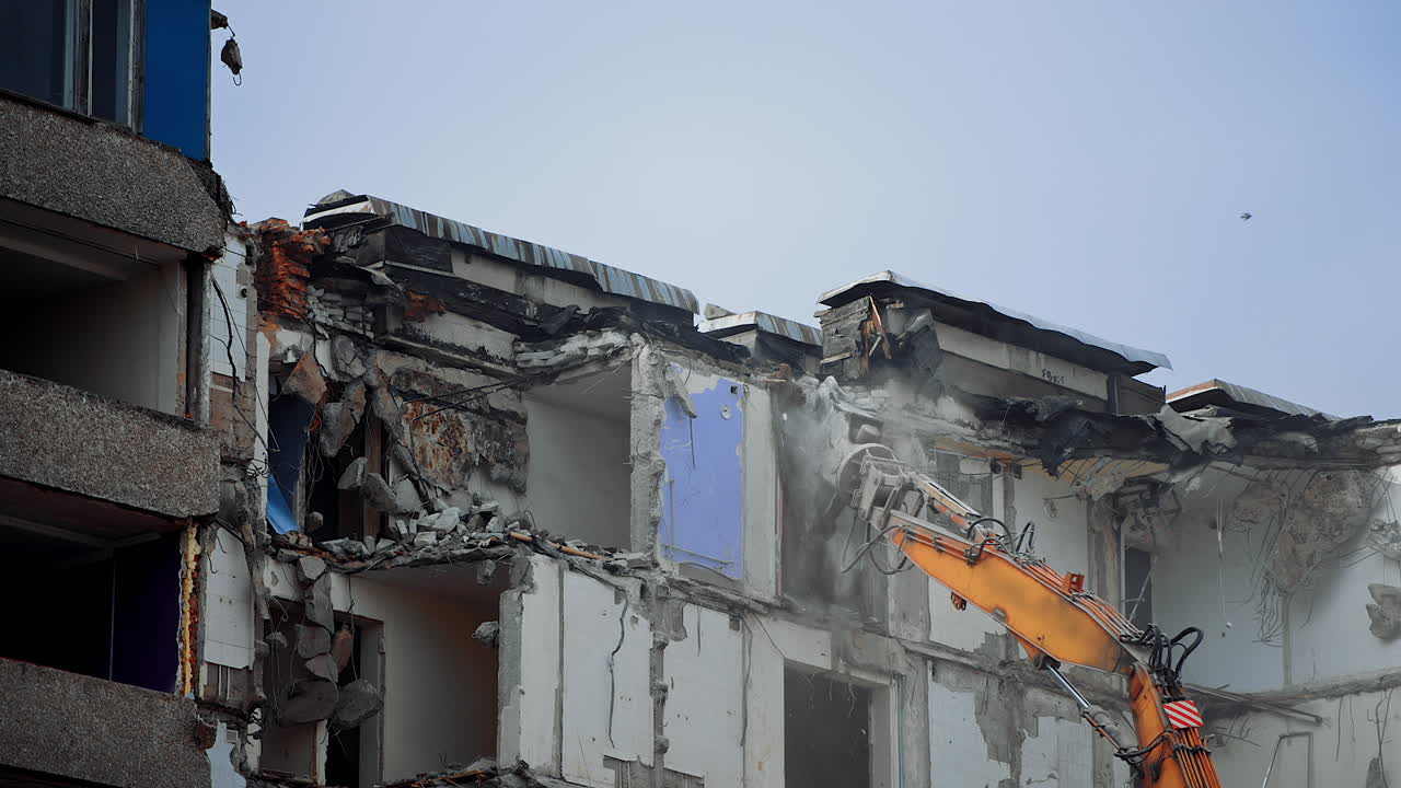 Excavator claw breaks the parts of ruined building. Dangerous deconstruction site with falling debris. Demolishing of the upper floors.