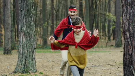 feliz padre caucásico y su pequeño hijo jugando a ser superhéroes con capas rojas en el bosque