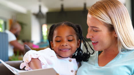 mother and daughter reading book at home together with family in background