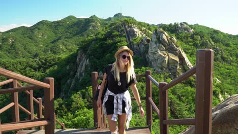 linda chica mochilera rubia en una caminata a la cumbre disfrutando del paisaje soleado, sosteniendo la barandilla y subiendo las escaleras para continuar el trekking