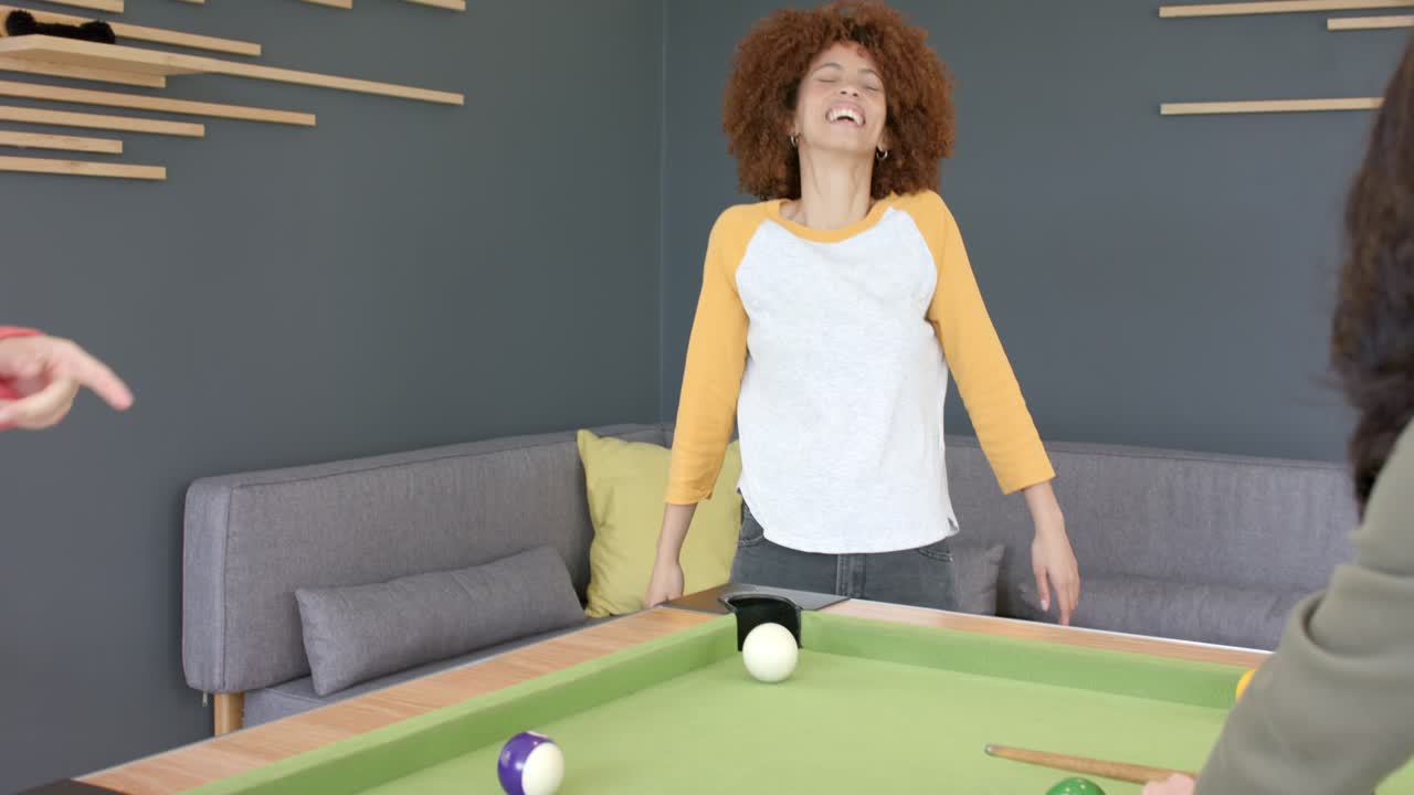 Diverse female friends noticing ball near pocket, blowing it on lounge pool table to sink ball