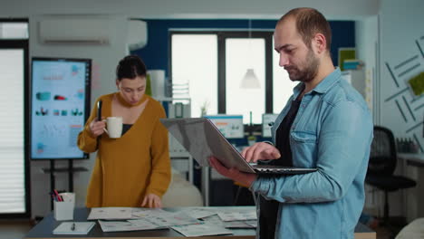 Portrait-of-casual-business-man-using-laptop-and-smiling-at-camera-while-coworker-holding-cup-looks-at-papers