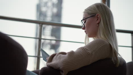 young lady seated, focused and writing on her journal with a thoughtful expression, wearing glasses, in a cozy indoor environment with natural light streaming through a glass panel