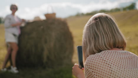 rear view of lady squatting taking photos of her sister in vast farmland with golden sunlight, scenic countryside setting with hay bale and picnic basket creating a warm rustic atmosphere