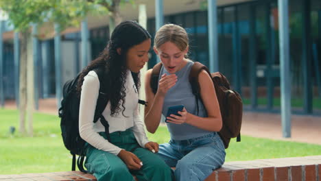 Two-Female-High-School-Or-Secondary-Students-Looking-At-Social-Media-On-Phone-Sitting-Outdoors