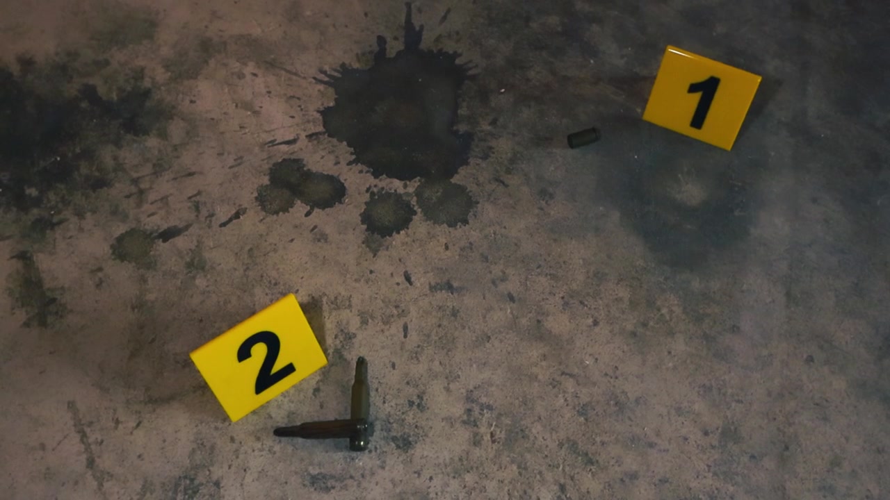 High caliber bullets and a 9mm casing lying next to numbered yellow evidence markers on a dusty and stained concrete floor, suggesting a recent criminal act and forensic investigation