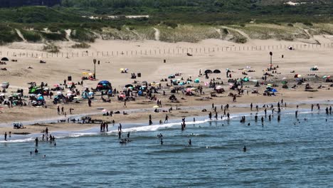 Multitudes-De-Personas-Disfrutan-De-Las-Vacaciones-De-Verano-En-La-Playa-De-Scheveningen,-Nadan-En-El-Mar-Y-Se-Broncean-En-La-Arena,-Holanda---Movimiento-De-Paralaje-Aéreo