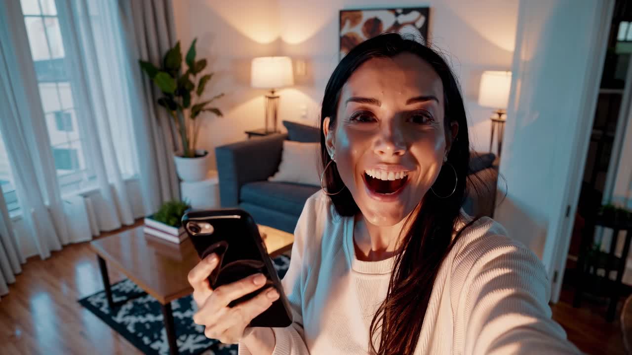 A woman smiles at the camera in a cozy living room, holding a phone