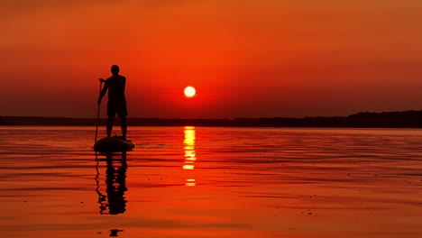 red sky and setting sun reflecting in the river. dark male silhouette approaching the camera on sup board.
