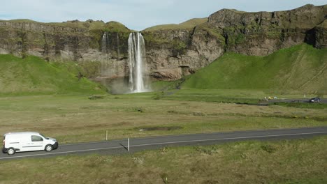 Weißes-Auto-Fährt-Auf-Landstraße-Mit-Wasserfall-Seljalandsfoss-Im-Hintergrund