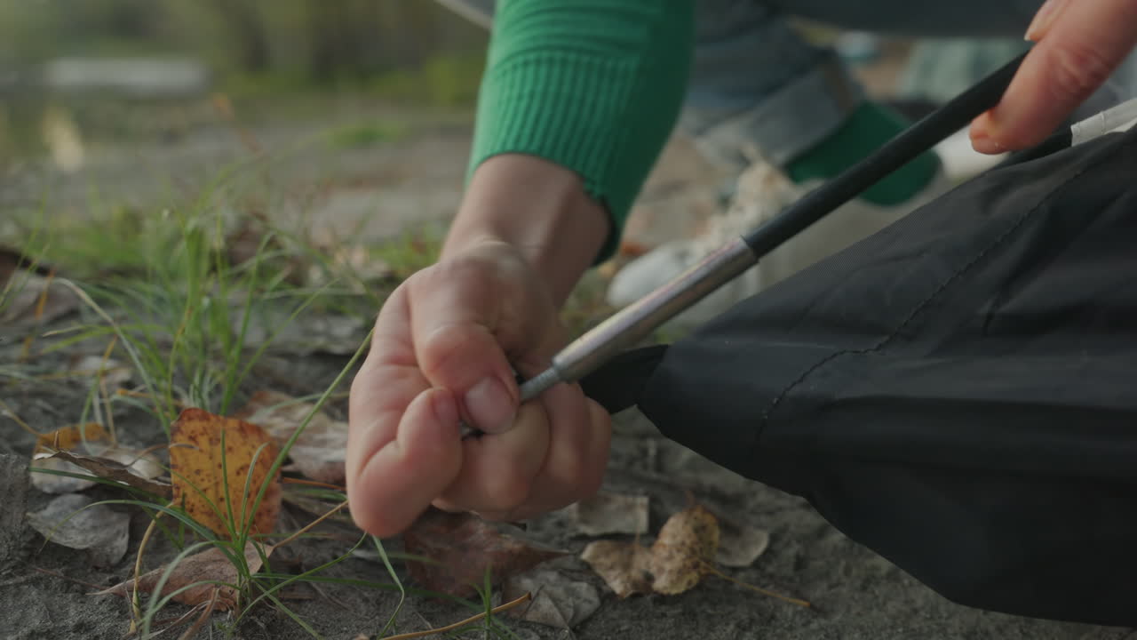 Closeup hands assembling tent pole, white woman securing stake on lakeshore, fabric tension against ground, fingers gripping metal pole, autumn leaves and evening light, focused camping setup