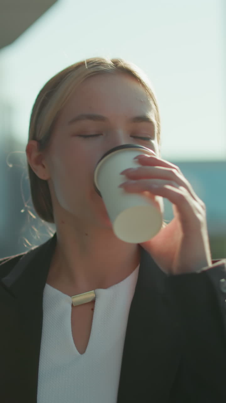 Woman with refreshing look in professional attire walks by modern glass building holding tablet in one hand and takeaway tea in other hand, sipping drink calmly in bright urban outdoor setting