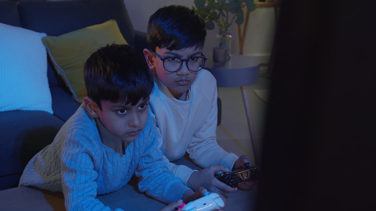Two Young Boys At Home Playing With Computer Games Console On TV Holding Controllers Late At Night 2