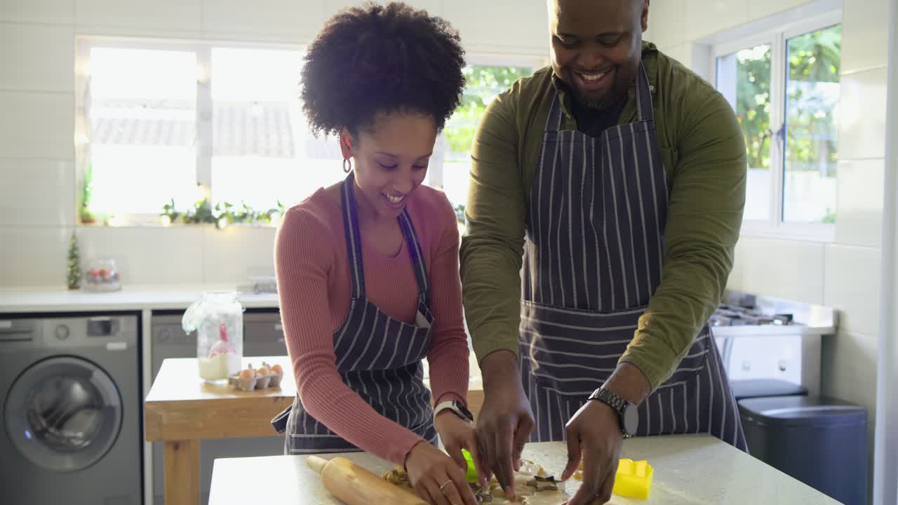 Diverse couple pressing holiday cutters into dough on home countertop and arranging cookies on tray