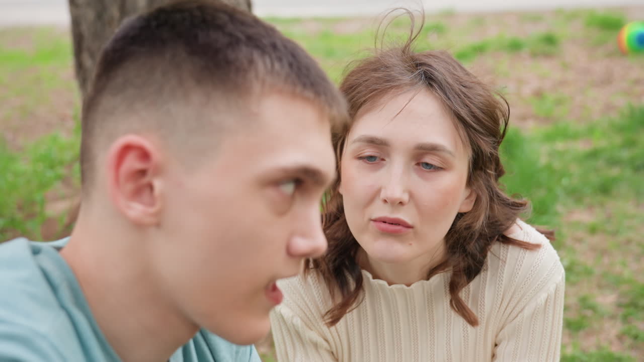 Young Caucasian Couple Sitting In Park Sharing Tense Conversation, Woman Listens With Concern While Man Speaks Animatedly, Closeup Faces, Tree And Grass Background, Candid Emotional Moment Blending