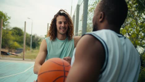 a curly red-haired man in a light t-shirt and a man with black skin in a white t-shirt are talking about basketball on the basketball court