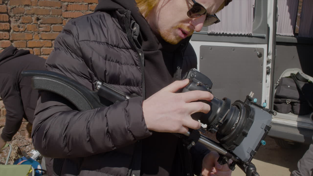 крупный план рыжеволосого работника производства в шерстяной шапочке и солнцезащитных очках, устанавливающего камеру на улице